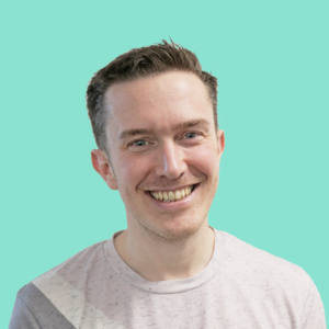 A headshot of Richard. He has short dark brown hair which it swept across to the left. He is smiling and wearing a pink, white and grey striped t-shirt.